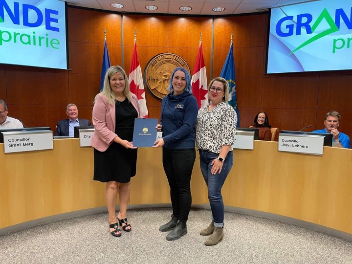 3 female presenting individuals smiling at the camera in front of city councillors holding a blue document folder